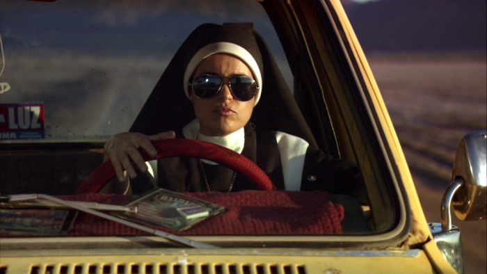 Screenshot from the film Symbol. A nun with sunglasses, played by Adriana Fricke, is driving an old yellow car in the Mexican desert.