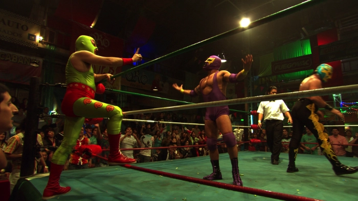 Screenshot from the film Symbol. An older Mexican lucha libre wrestler in green and red tights wags his index finger while he's standing outside the wrestling ring. The other wrestler in purple tights has his hands in the air as he challenges the green wrestler.